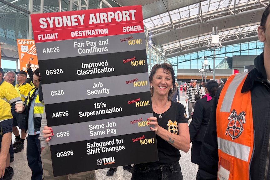 A woman with short hair smiles holding a large sign demanding better working conditions for airport ground crew.