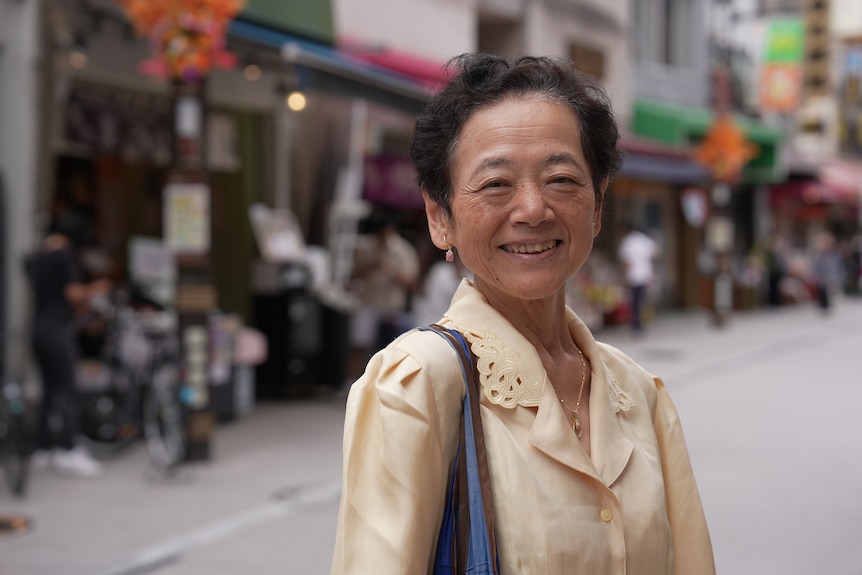 A smiling Japanese woman wearing a creme yellow blouse while standing in an empty roadway.