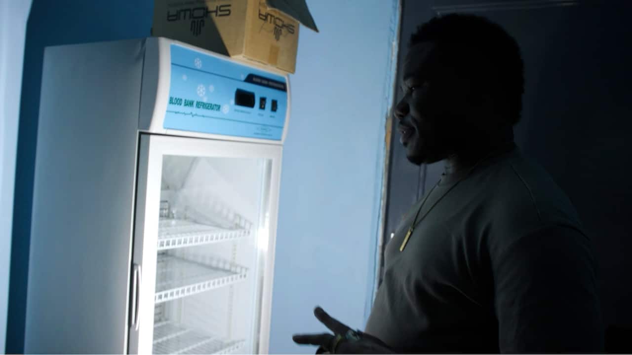 A man stands beside an illuminated empty blood bank refrigerator in a dark room. 