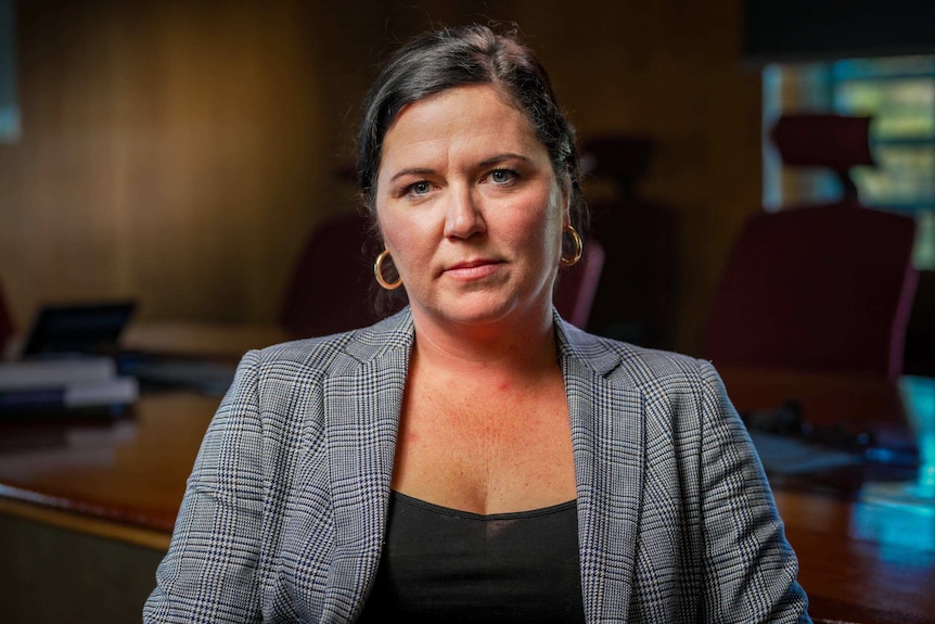 A woman wearing a grey check blazer and black top sits in a boardroom.