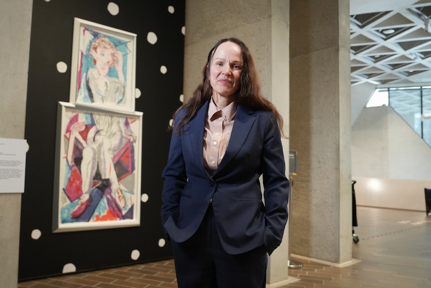 A woman with long brown hair wearing a navy suit stands in front of a neo-cubist color lithograph on the wall of an art gallery.