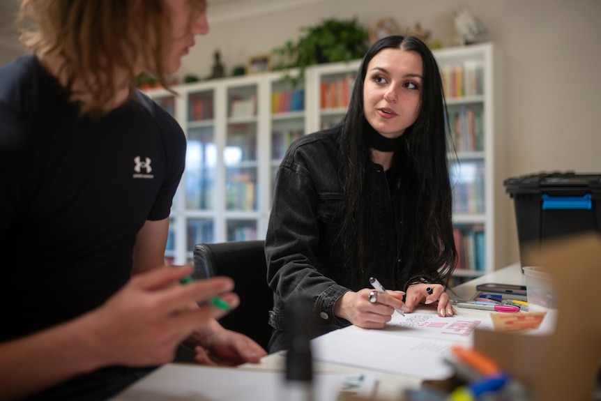 Three young people create colourful posters protesting AI data centres with book shelves in the background.
