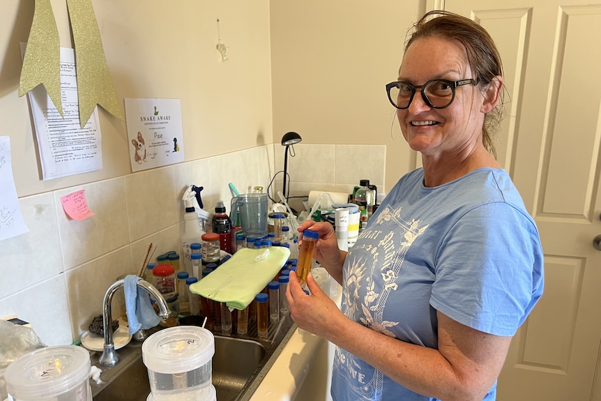 A woman stands next to a sink with many jars and tubes filled with liquid.