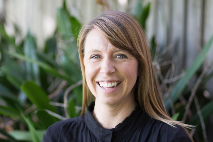 Bel, a woman with straight hair, wears a black top and smiles as she sits in front of a leafy backdrop.