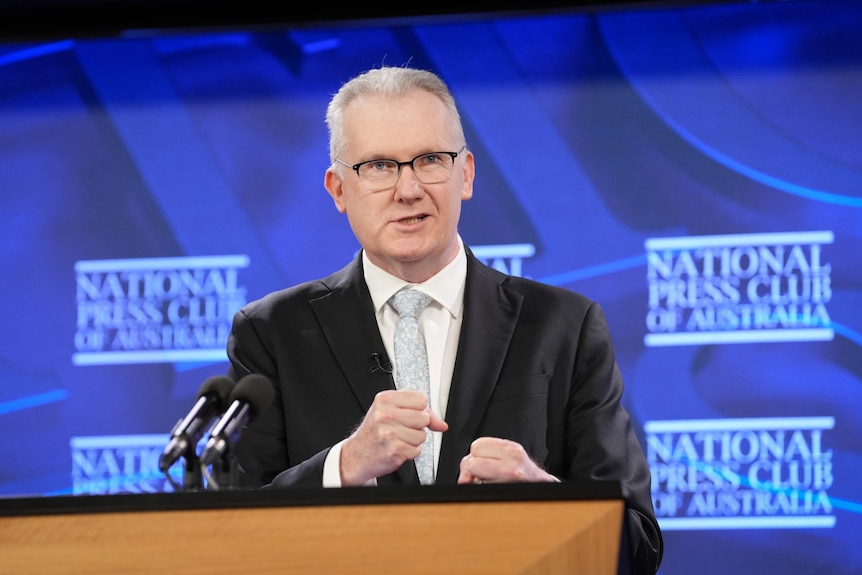A man in a suit stands at a podium talking to a crowd.
