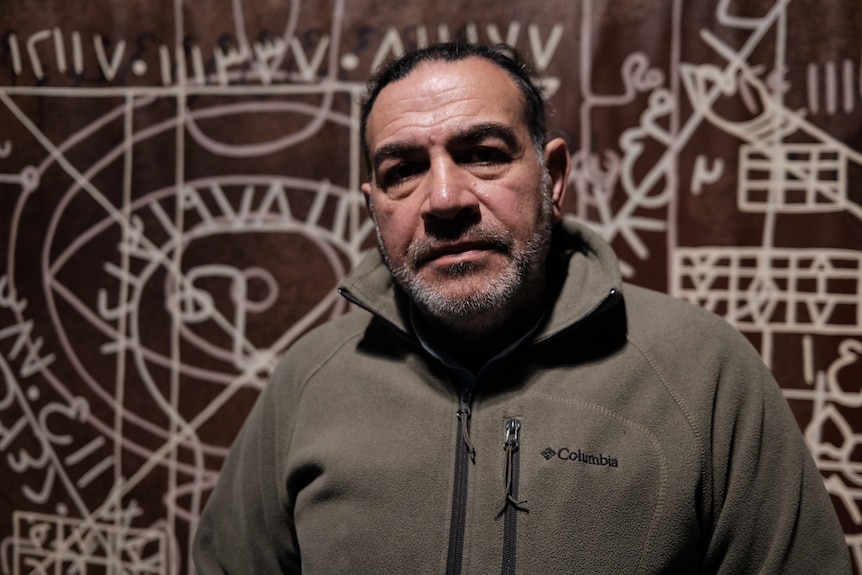 A bearded dark-haired man (Khaled Sabsabi) stands in front of some brown artwork.
