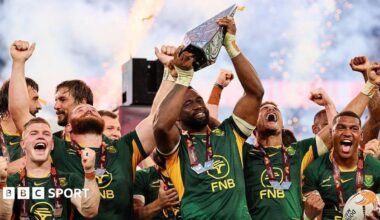 Siya Kolisi lifts the Rugby's Greatest Rivalry trophy, surrounded by happy team-mates in South Africa's largely green kit, as pyrotechnics light up the background