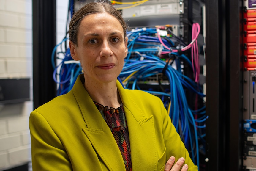A woman with hair tied back wearing a green blazer stands in front of technical equipment and computer cords