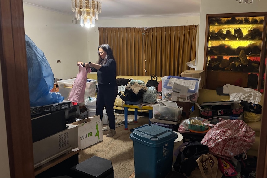 Woman folds clothes in a messy lounge room. Wide view of the room shows her using couch as table for basket.