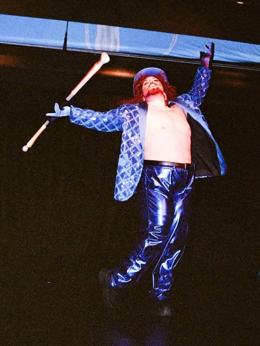 A drag queen in a purple reflective suit and top hat twirls a stick on stage.