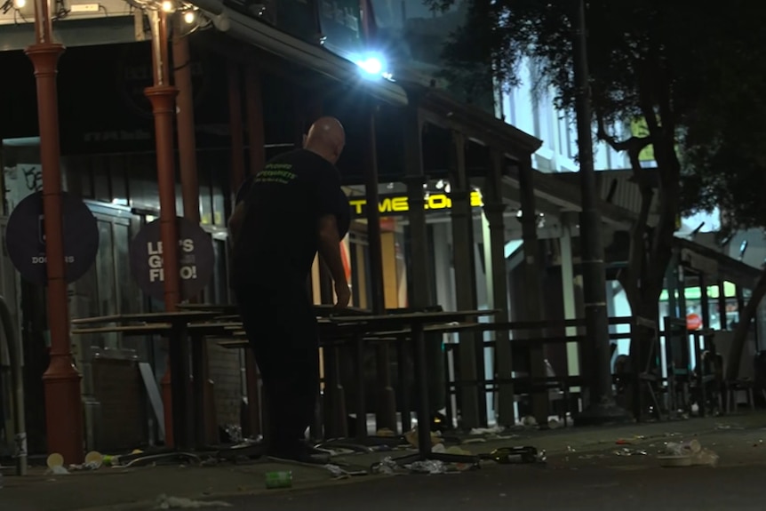 A worker moves a table off a footpath covered in rubbish outside a hotel at night.