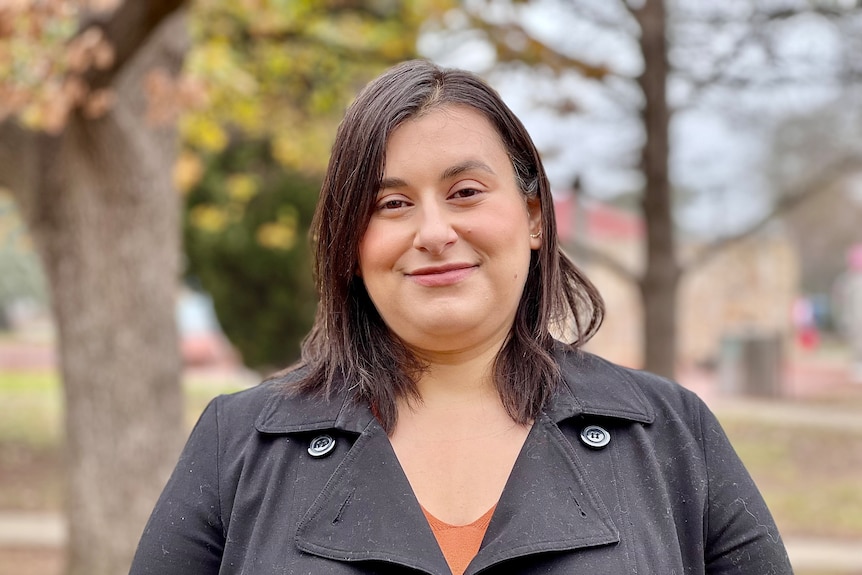 A woman with shoulder-length brown hair, wearing a black jacket and orange shirt, smiles outdoors.