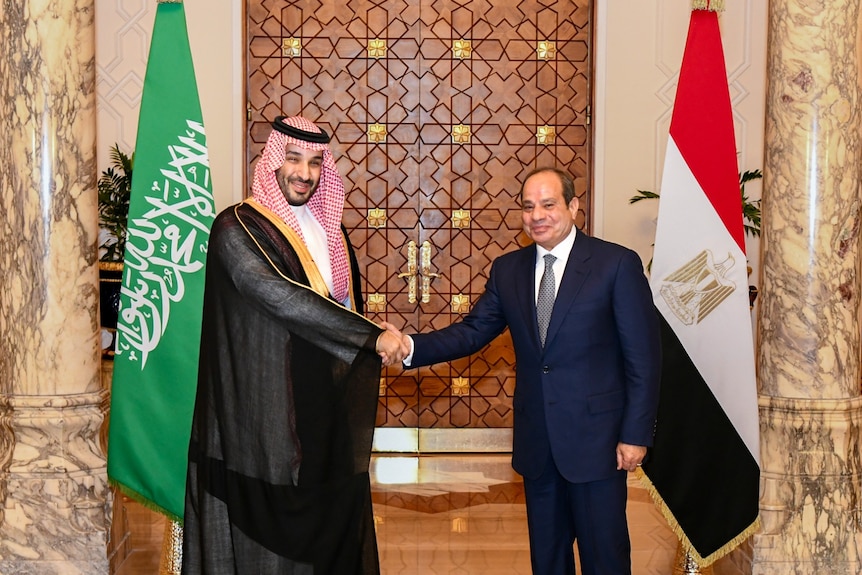 Crown prince in black robes, red and white headdress shakes hands with a man in suit, both smiling, in front of national flags.