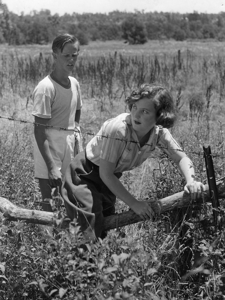 Black and white photo from the 1940s or 1950s showing a teenage boy and girl climbing through a fence on a rural property
