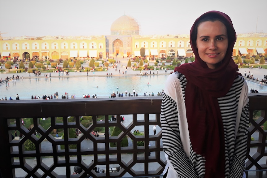 A young woman wearing a headscarf stands in front of a large courtyard and mosque.