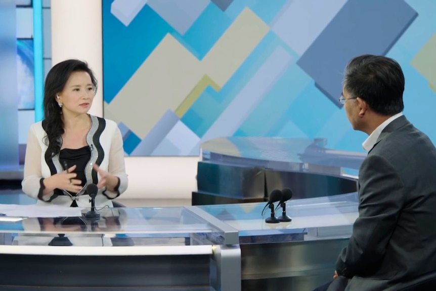 A female news reporter seated at a television news set desk across from a man she is interviewing.