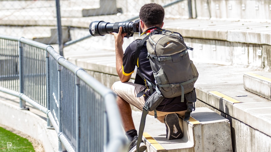 Photographer sits on concrete stadium seats with his back to the camera and aims a camera lens out over a field with a backpack