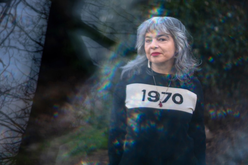 A woman with silver hair is seen through a glass prism in a jumper with "1970" printed on the front.
