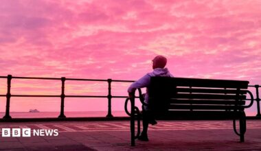 A man in a grey hoody and beanie with glasses is sat on a bench faced away from the camera. He is looking out at the sea and a boat. The sky is full of a beautiful pink hue with yellow and purple tints.
