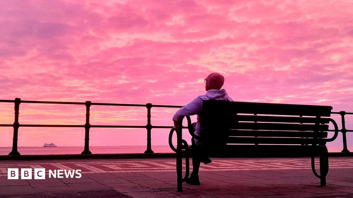 A man in a grey hoody and beanie with glasses is sat on a bench faced away from the camera. He is looking out at the sea and a boat. The sky is full of a beautiful pink hue with yellow and purple tints.