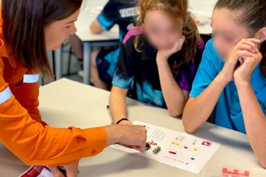 A woman in high-vis explains a worksheet to school students sat at a desk.