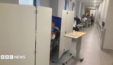Image shows a hospital corridor. There is a bed on the left-hand side of the image, surrounded by privacy screens. An elderly man is just visible lying on a bed largely obscured behind the screens. In the background, a number of other, mainly elderly, people can be seen on beds and trolleys lining the wall of the corridor.