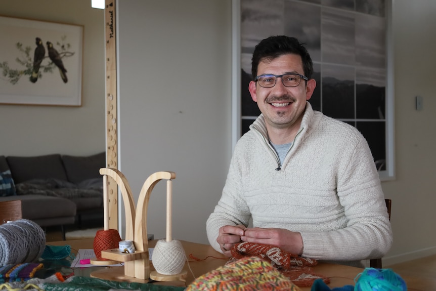 Will Seager sits at his dining room table upon which are several finished projects, and holds a shawl he is knitting.