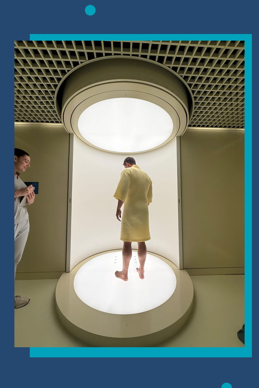 A person in a yellow robe stands on a circular platform under a bright light, while another person observes nearby.