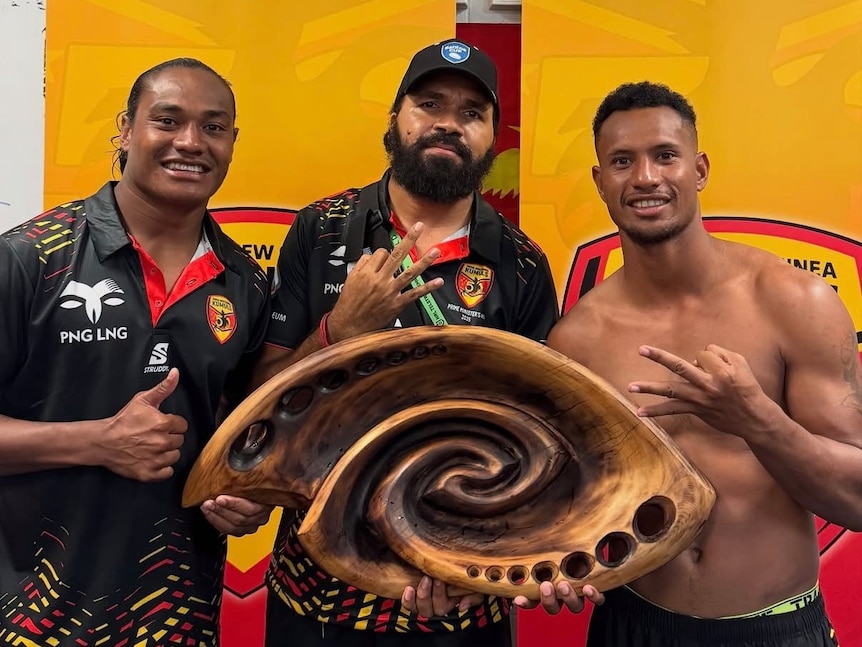 Three PNG men, two wearing black gold shirts and one with no shirt, pose with a wooden trophy and smile at the camera.