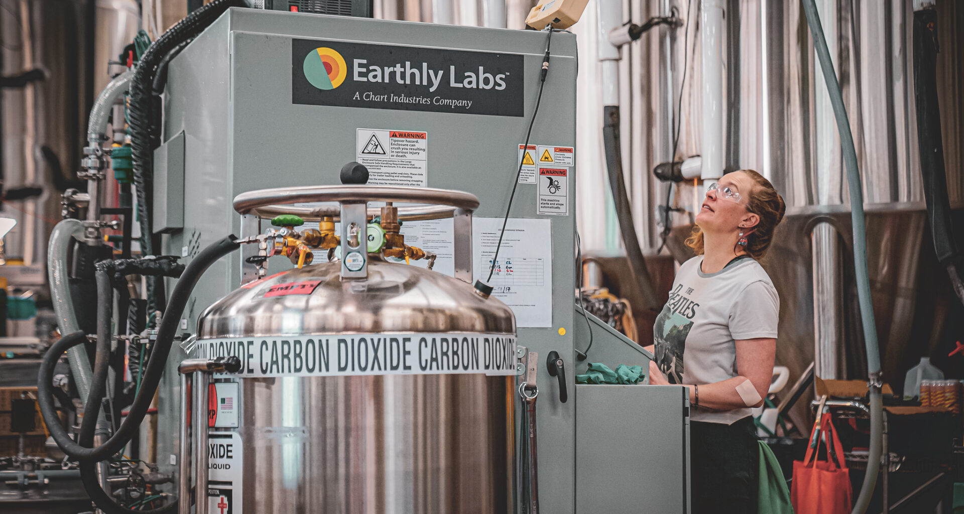 A woman controls an Earthly Labs-branded machine in a factory setting; a tank that says “carbon dioxide” is in the foreground.