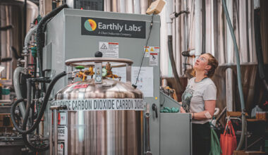 A woman controls an Earthly Labs-branded machine in a factory setting; a tank that says “carbon dioxide” is in the foreground.