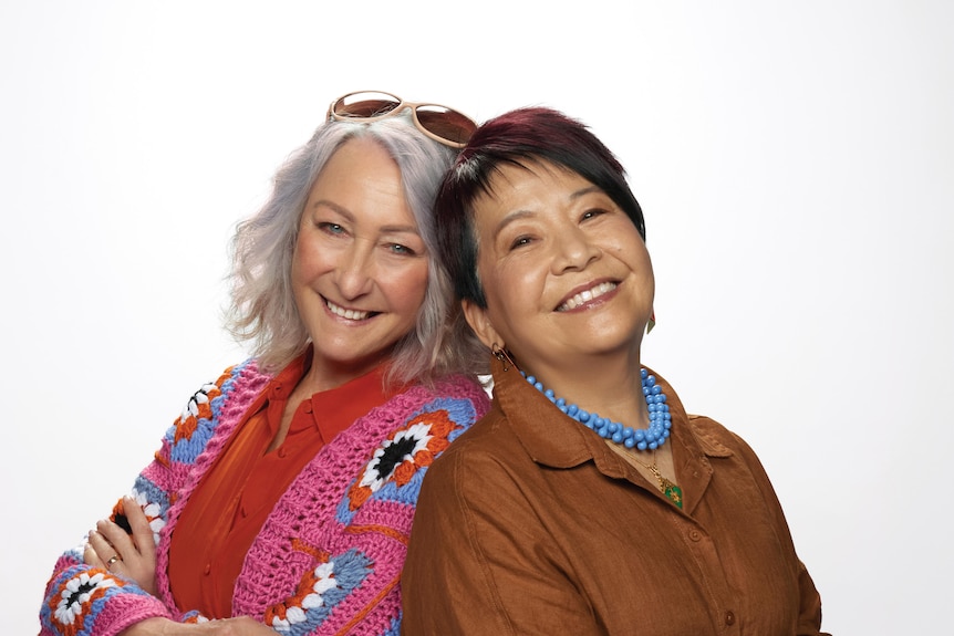 Lynne McGranger, 73, in a pink cardigan, and Annette Shun Wah, 68, in blue beaded necklace, pose back to back, grinning widely.