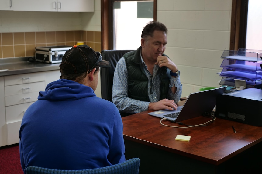 A man in a blue hoodie and a black cap sits with his back to the camera. Another man taps on his computer behind him.