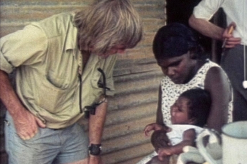 A man in a beige shirt leans down to talk to an Indigenous woman cradling a baby.