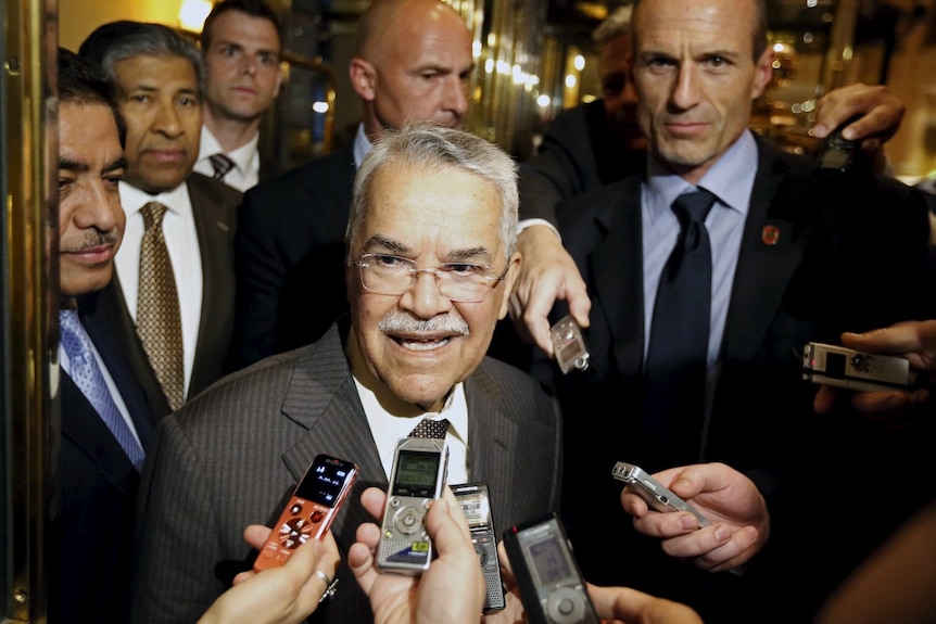 A man in a suit speaking, surrounded by microphones and other men.