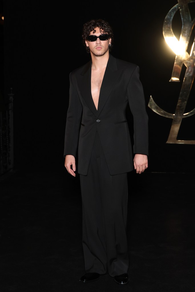 Belmont Cameli wearing a black suit with no shirt underneath and sunglasses at the Saint Laurent show during Paris Fashion Week.