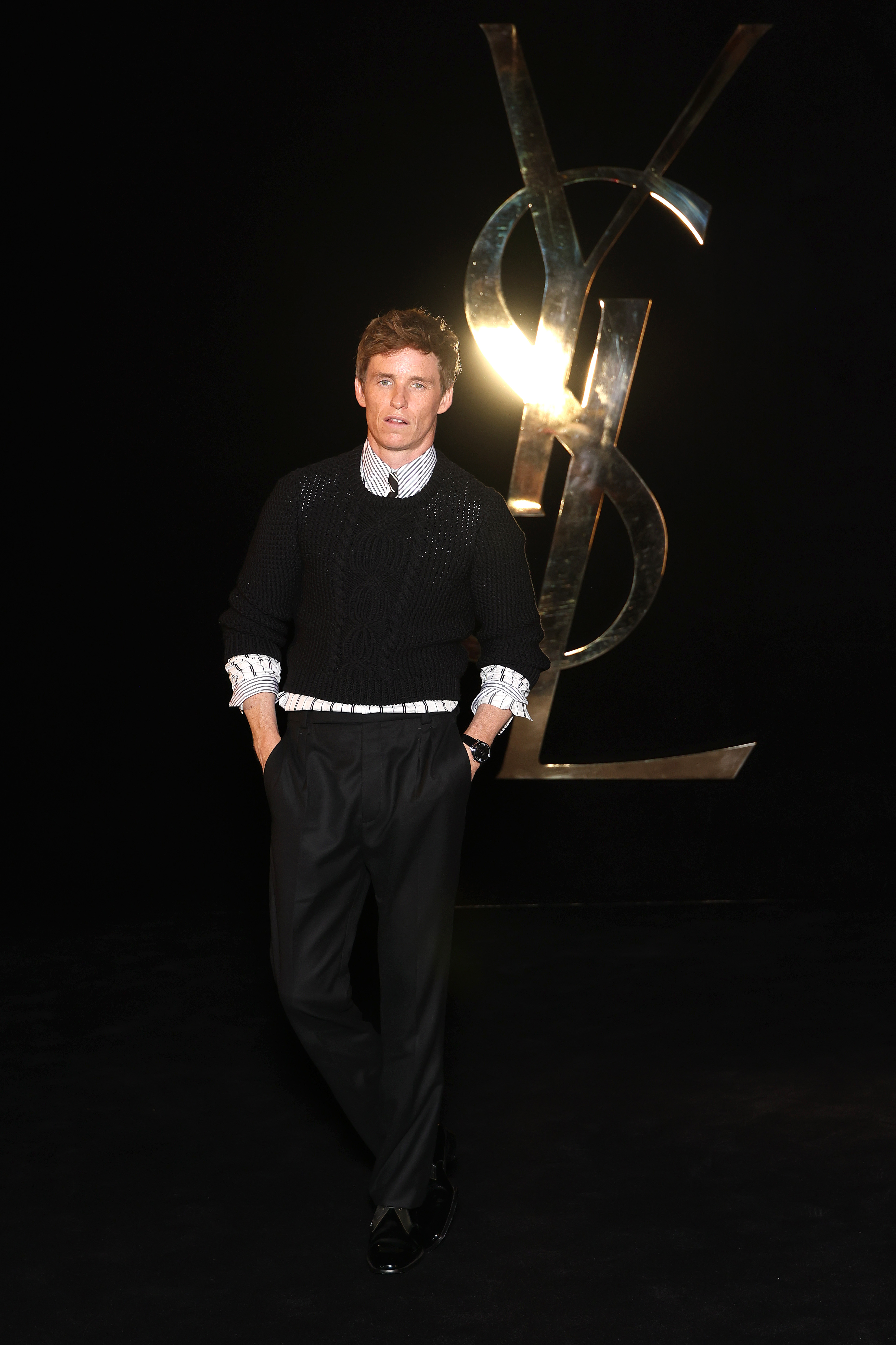 Eddie Redmayne poses in a black sweater over a striped shirt and black trousers, with a large illuminated Saint Laurent logo in the background.
