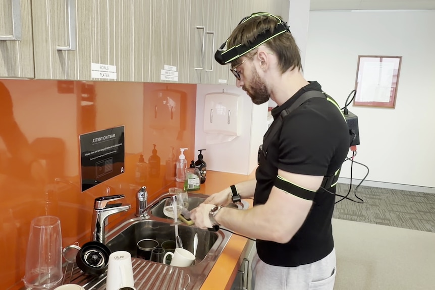 A man washes dishes wearing an a camera and sensors on his hands.