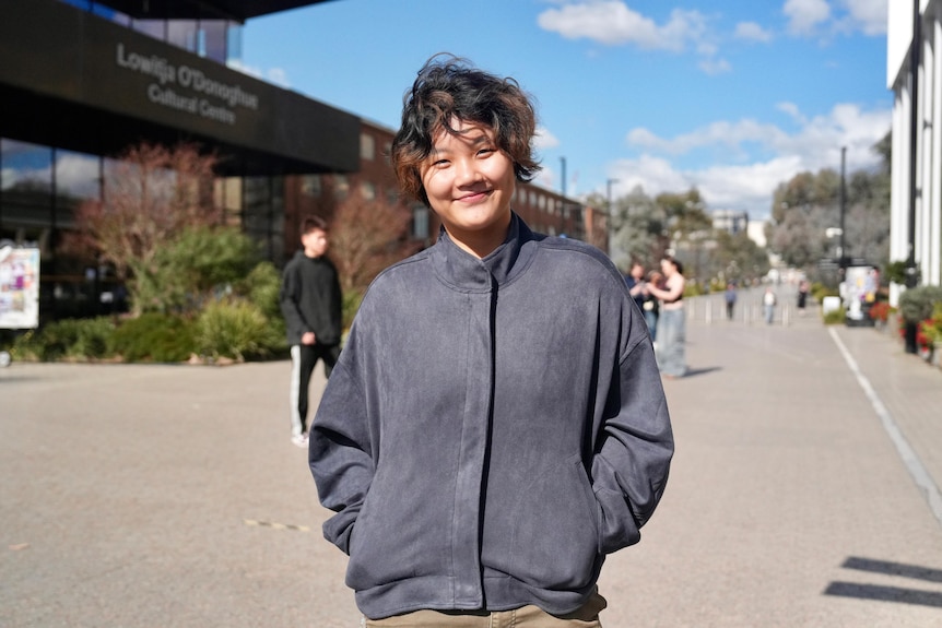A woman of Asian appearance with short curly hair and hands in pockets smiles and stands on walkway.