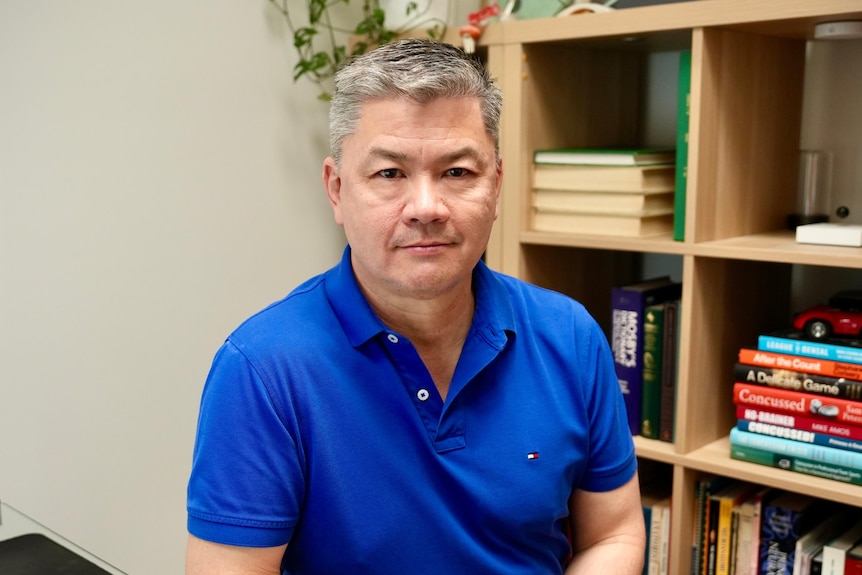 A man in a polo shirt sits in an office, looking into camera with a serious expression.