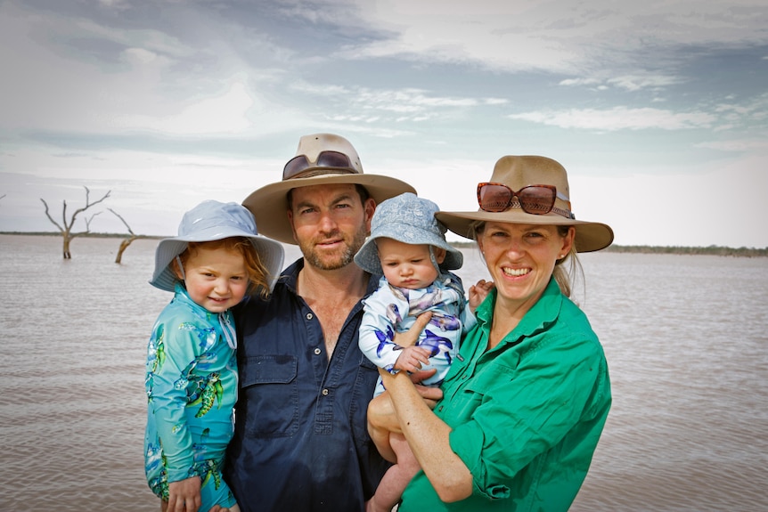 A man and a woman wearing wide-brimmed hats hold a baby boy and toddler girl while standing at a large dam.