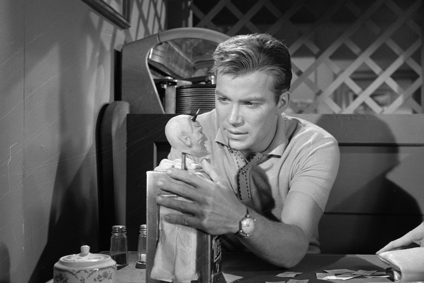 William Shatner holds a fortune-telling machine in black and white still from a TV show