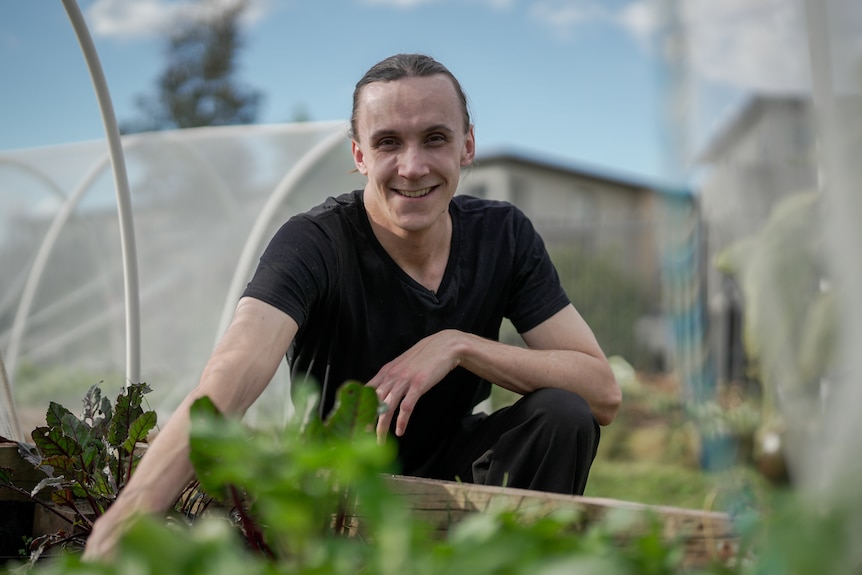 A man wearing a v-neck black shirt with his hair tied back squats next to a garden bed with green seedlings in it.