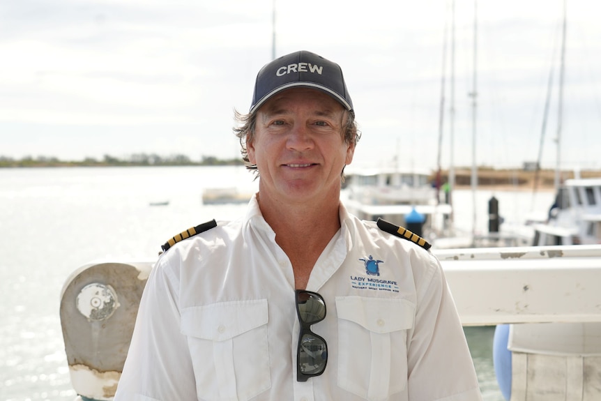 A man in a ship's captain white shirt with sunglasses in his pocket, standing in front of boats at a marina.