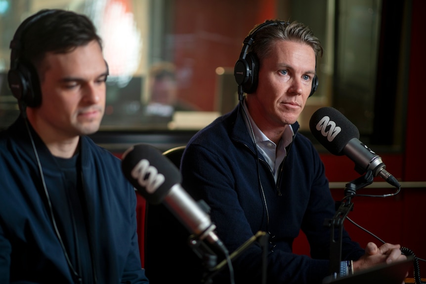 Two young men sit in a radio booth with red walls, wearing headphones and speaking into an ABC branded microphone.