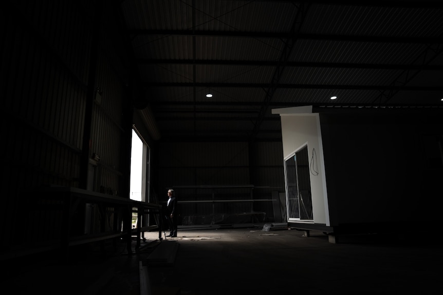 A man stands at the door of a warehouse with a partially finished home behind him
