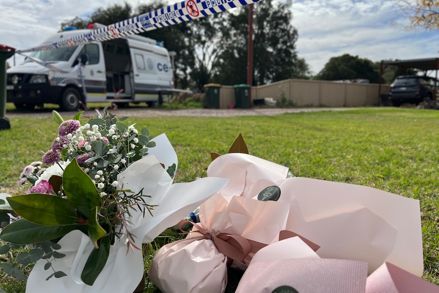 Flowers left outside a house with police tape above the flowers and a van in the background.