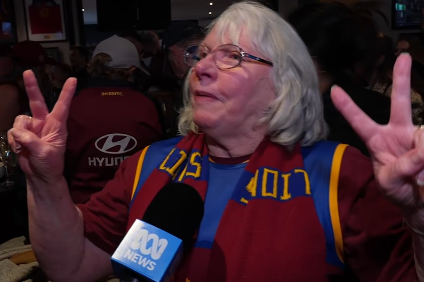 A white haired woman wearing glasses and a Brisbane Lions scarf holds her two hands up with two fingers up.