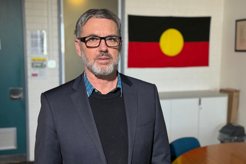 Mr Newhouse in a blazer and dark blue jumper standing in a room looking serious with an Aboriginal flag behind him.