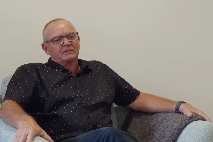 A balding man in a dark shirt seated in an armchair